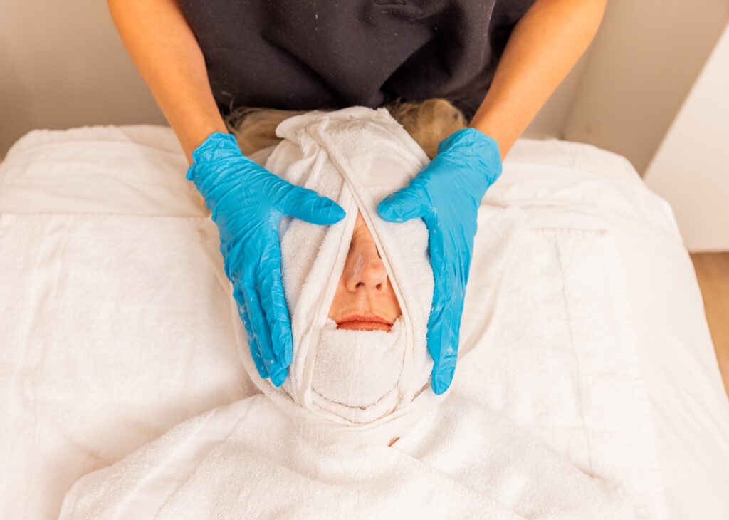 A patient at a med spa lying in a treatment bed while gloved hands wrap a towel around their face for administering one of Ona Skincare's best Chemical Peels Near Belle Meade.