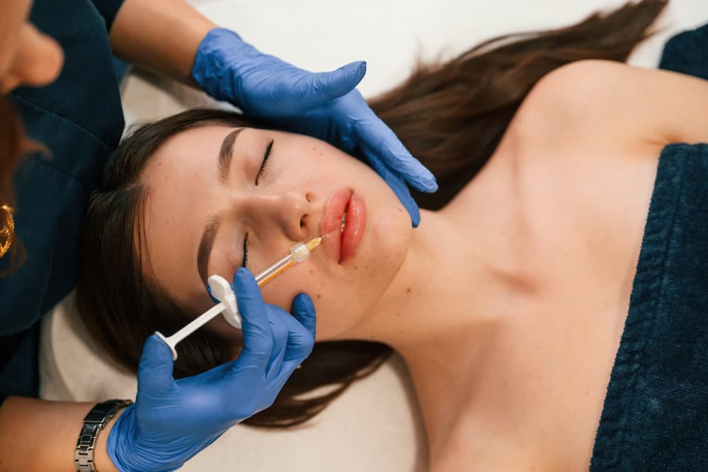 A young woman lying down, receiving lip filler near Belle Meade from a woman wearing gloves.